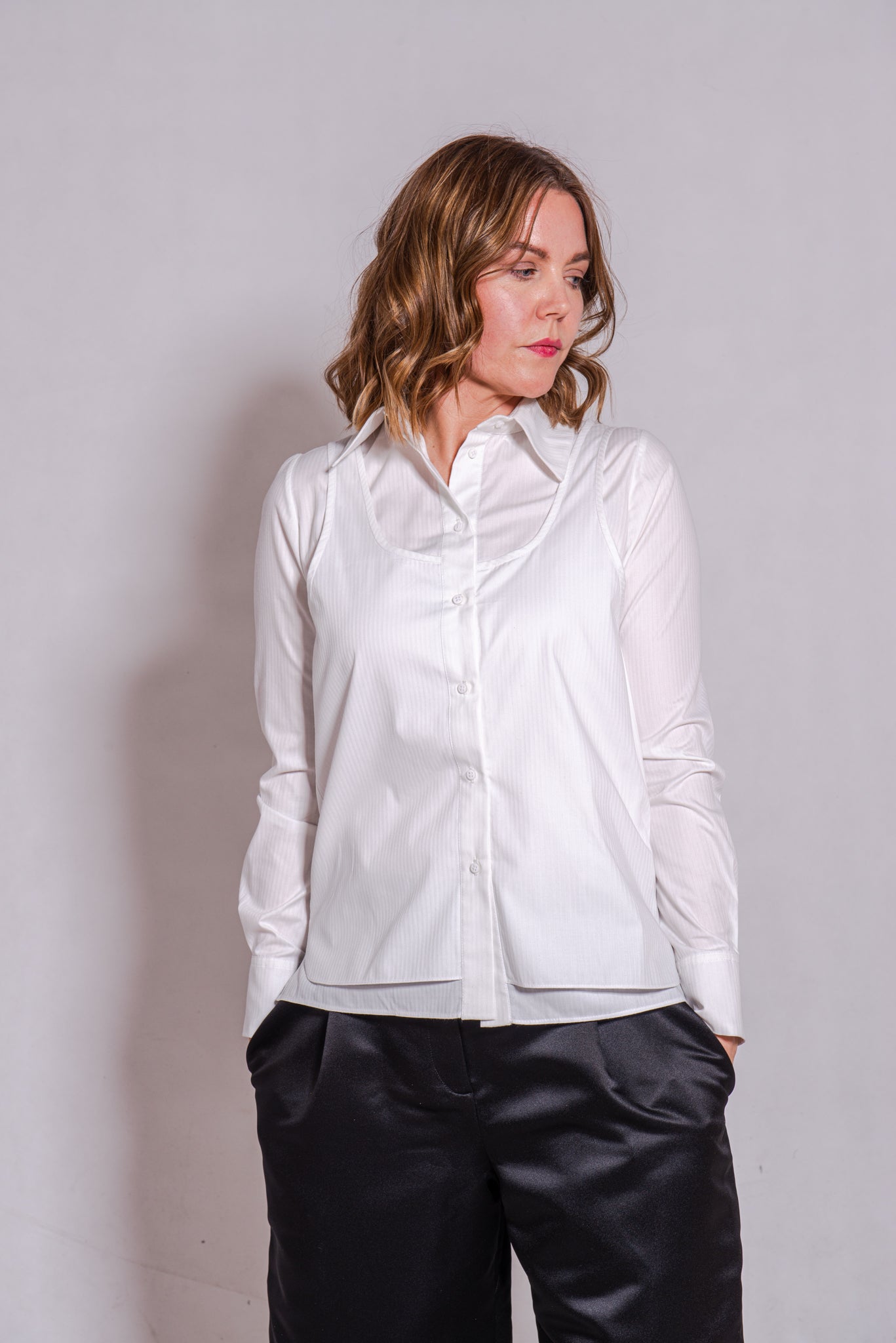 Women wearing Abbey shirt white paired with Agnes trousers (black) against a grey wall.