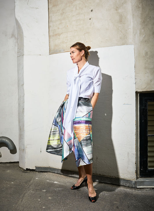 Model standing by port entrance wears white Alan short sleeved shirt paired with an Alayanvskirt (multi)