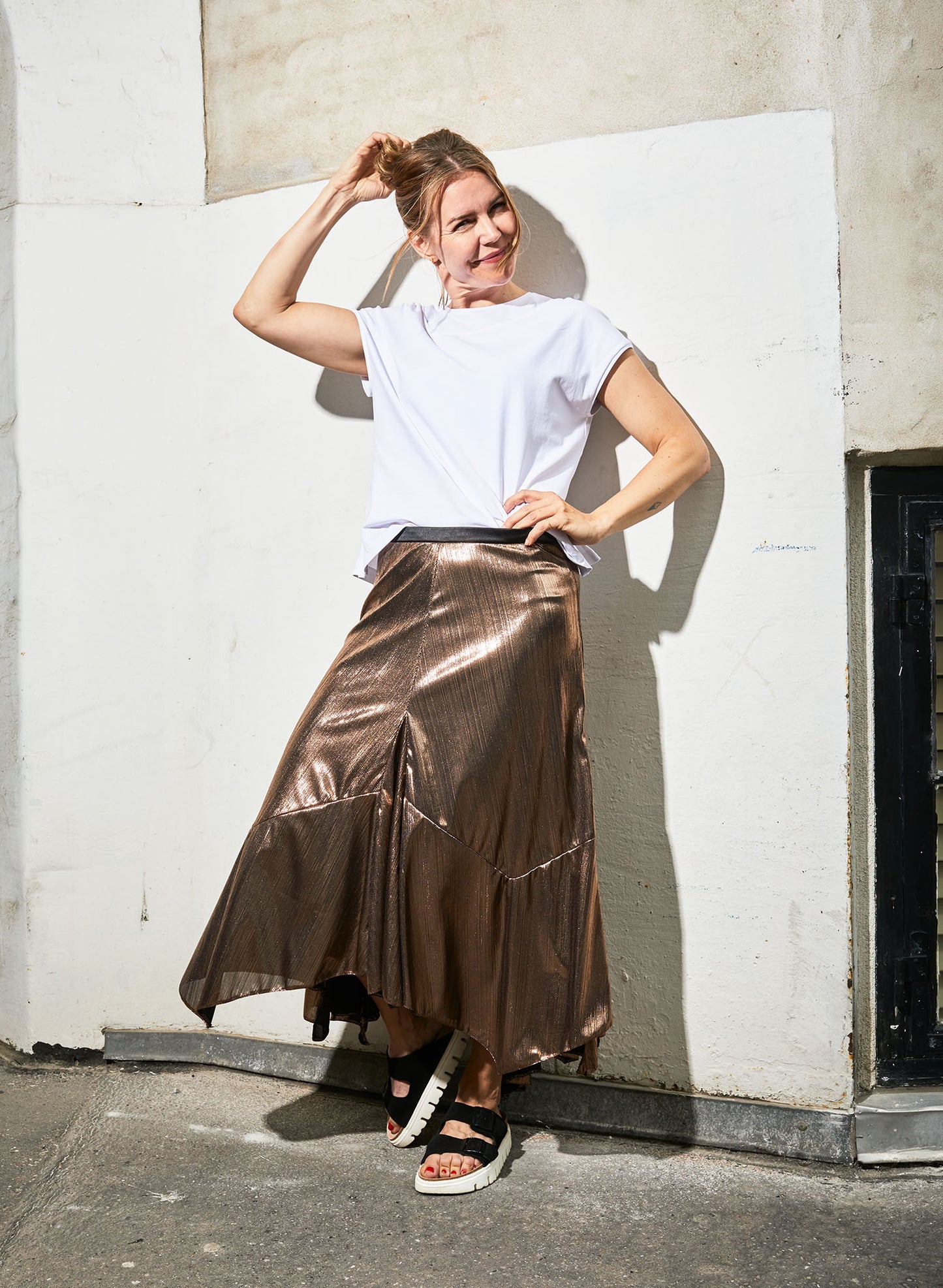 Model, in front of geometric lined wall, wearing Alyza skirt bronze paired with models white tshirt