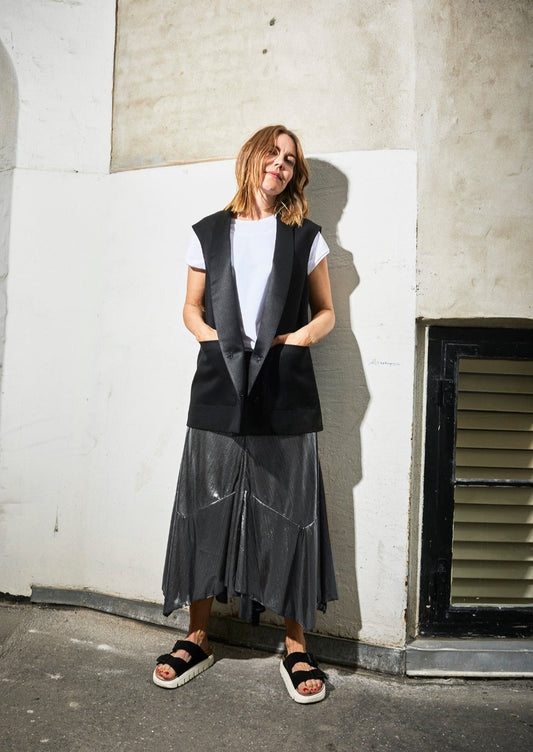 Model smiles, in front of geometric lined wall, wearing Alyza skirt, pewter, paired with Alison Gilet black & with models white tshirt