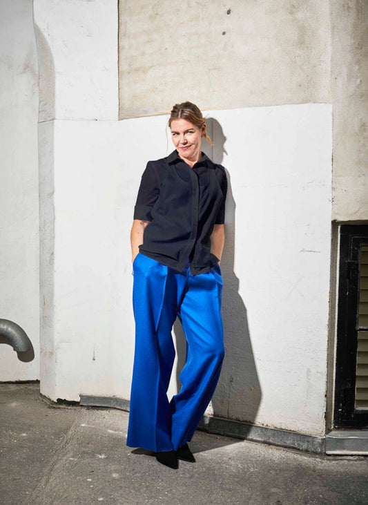 Women wearing Abba shirt white paired with Ava pants (blue wool) leaning against a black wall