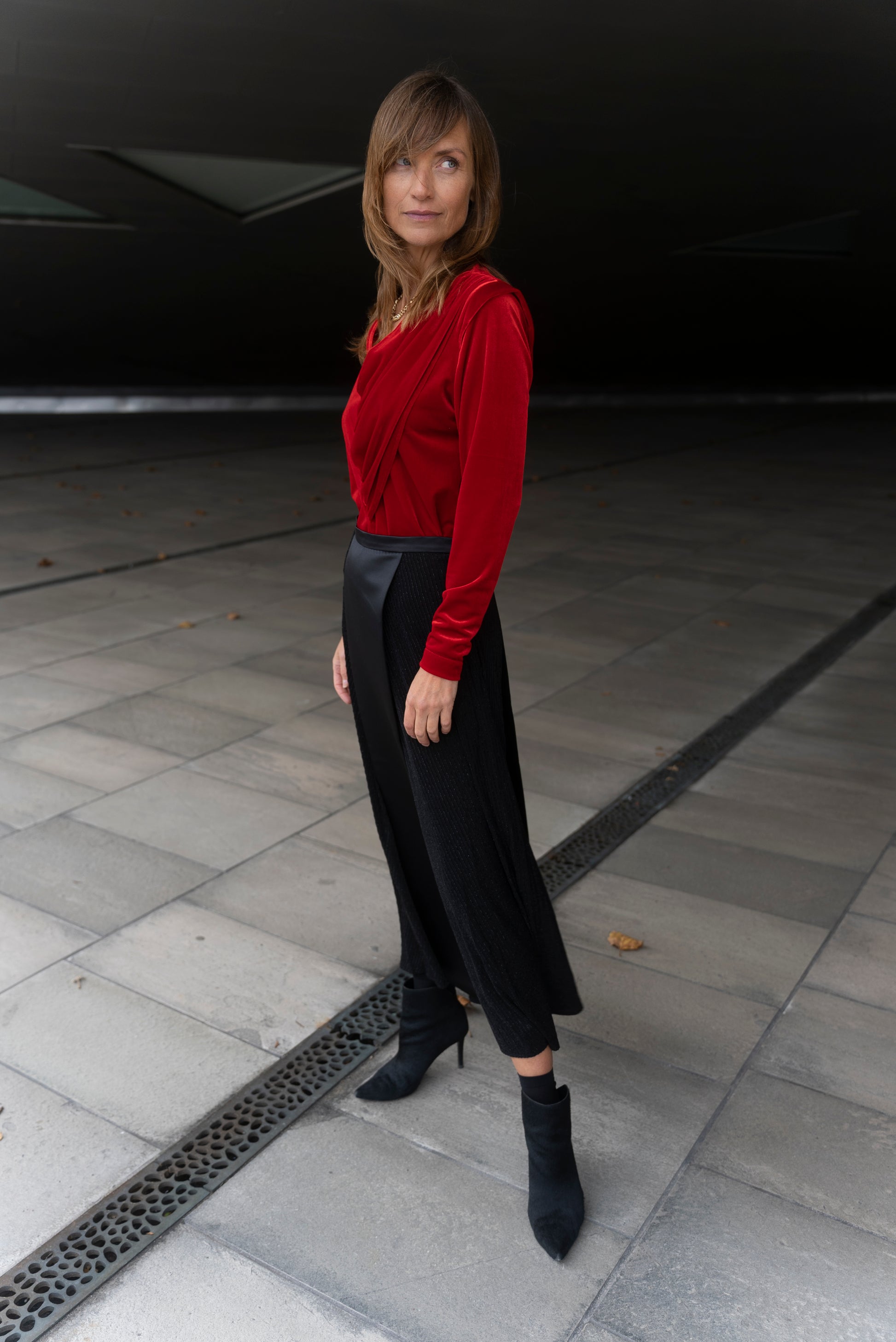 Model wearing Alek top red velvet and Aya skirt stripey black full length. Under geometric building.