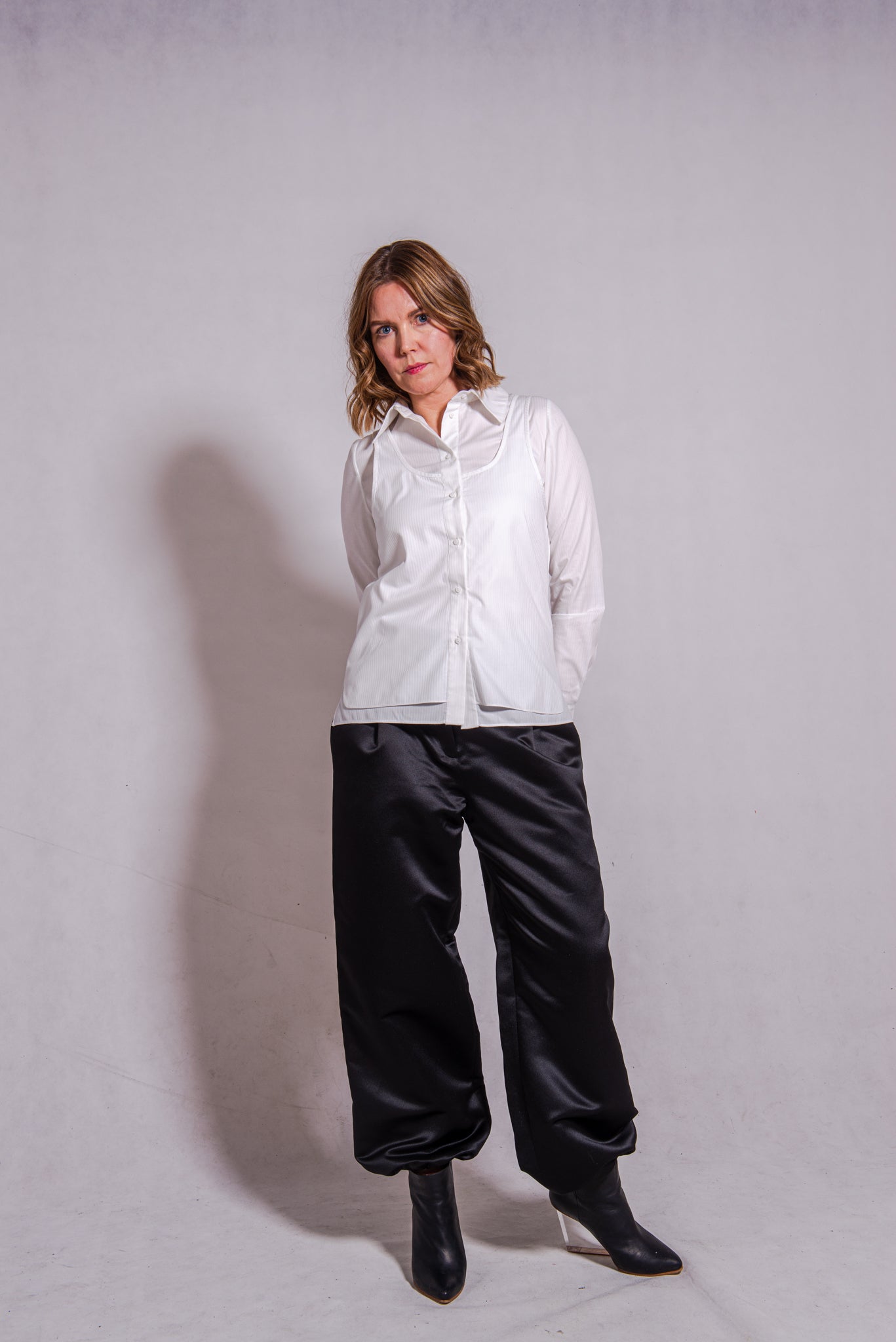 Women wearing Abbey shirt white paired with Agnes trousers (black) against a grey wall.