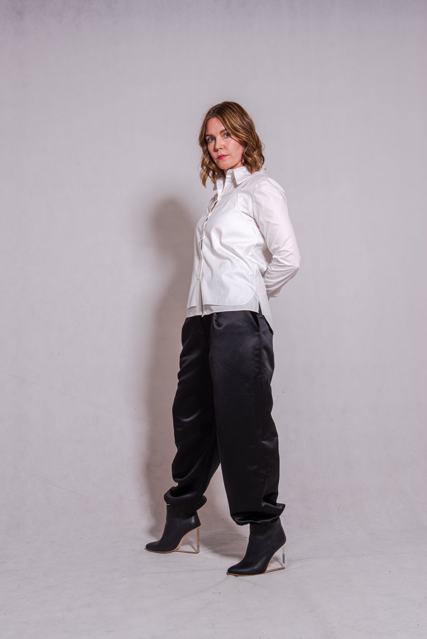 Women wearing Abbey shirt white paired with Agnes trousers (black) against a grey wall.