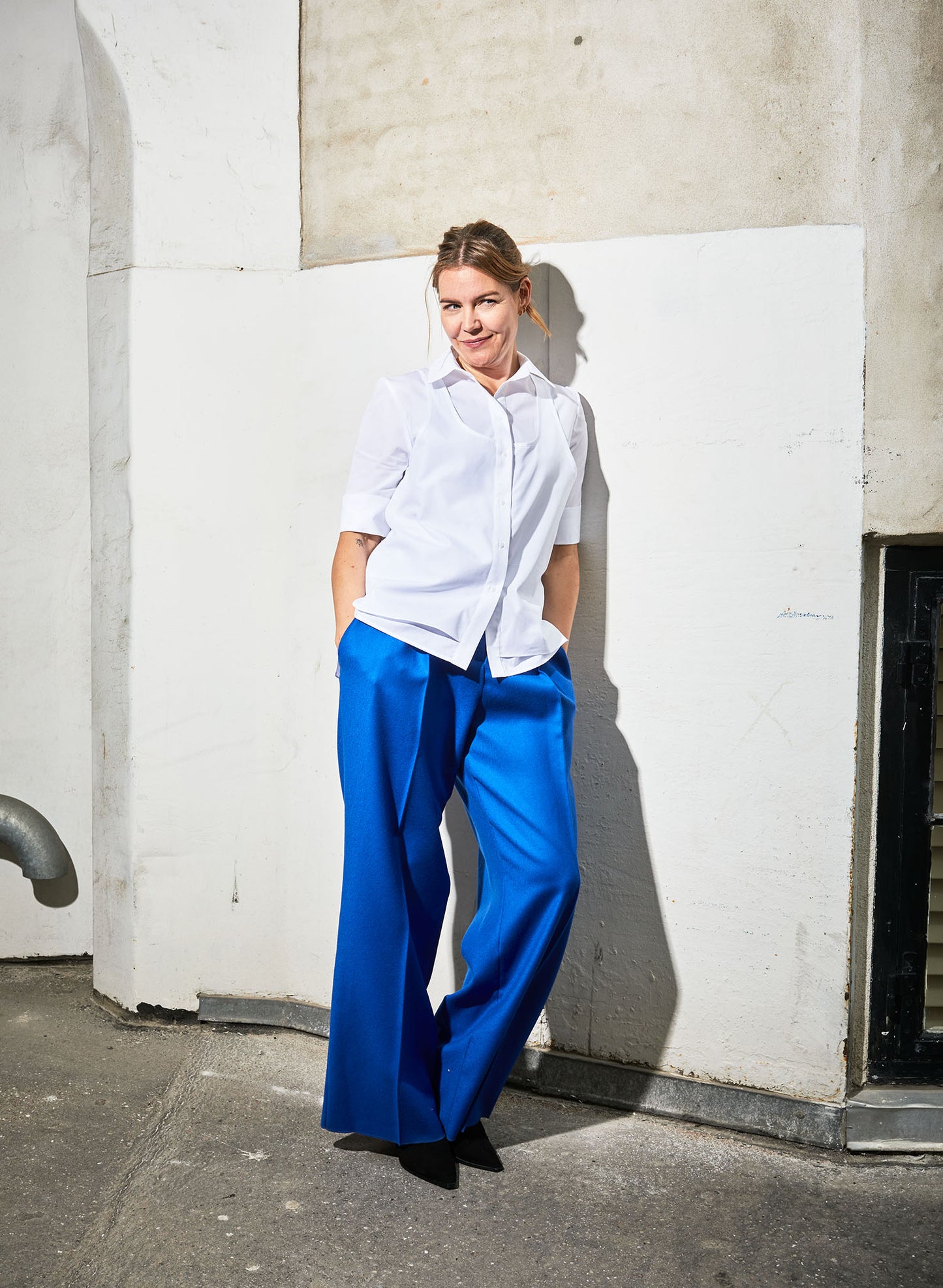 Women wearing Abba shirt white paired with Ava pants (blue wool) leaning against a white wall.